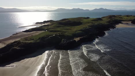 establishing aerial view ynys llanddwyn welsh island with shimmering ocean and misty snowdonia mountain range across the sunrise skyline