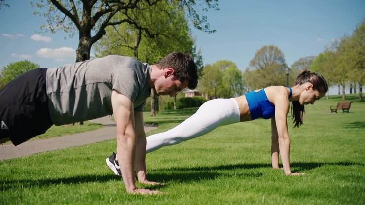Holding sunlight-prompted man and woman maintaining high plank on grass in park, wearing sportswear