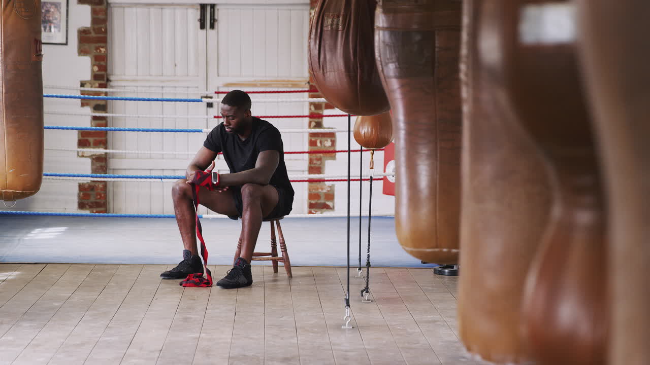 Male Boxer Training In Gym Putting Wraps On Hands Sitting Next To Boxing Ring And Punching Bags