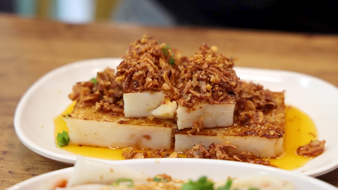 Chopsticks pick up a piece of fried turnip cake topped with crispy shallots from a white plate in a brightly lit Singapore eatery, captured in a medium close-up
