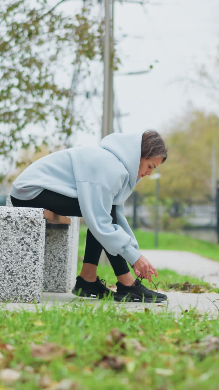 Young woman sitting on concrete bench tying her shoelace, with blurred background showing trees, iron fence, grass, and pole in an outdoor park, preparing for exercise or walk