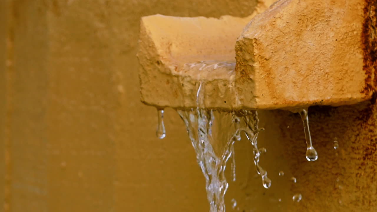 Water Flowing from a Stone Gutter on a Textured Wall, Stream of Water Pouring from a Stone Spout, Water Cascading from a Concrete Channel, Water Dripping from a Rough Stone Gutter.