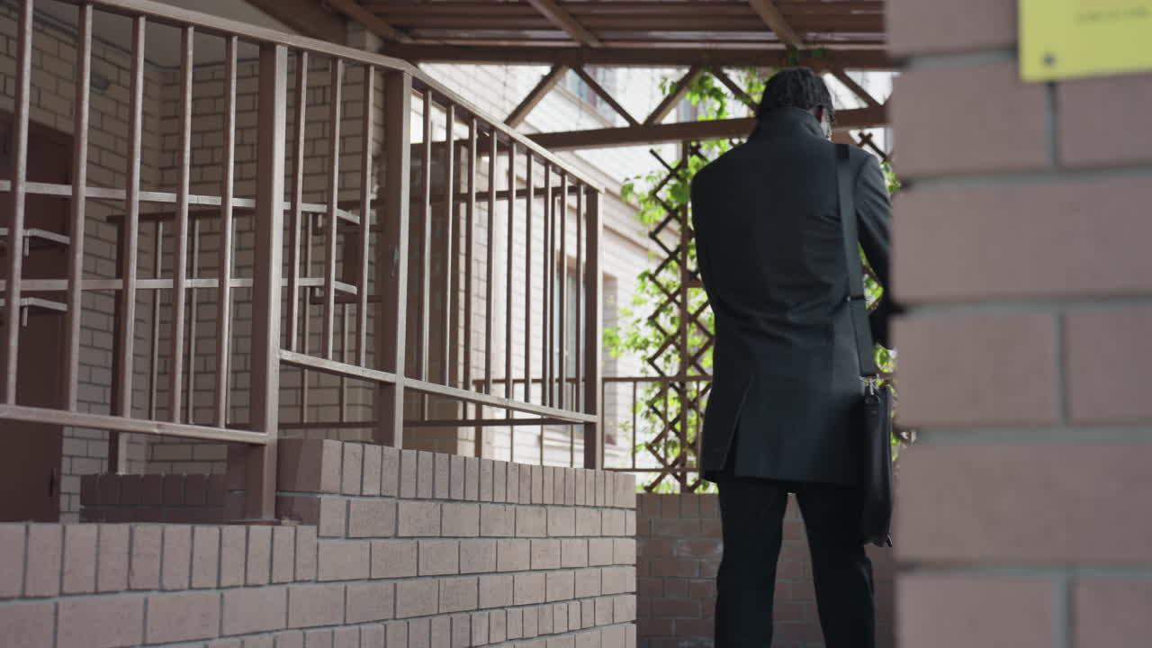 rear view of adult with natural Locs in dark overcoat climbs outdoor steps toward balcony, shoulder bag strap visible, brick corridor, iron railings with soft daylight create candid urban vibe