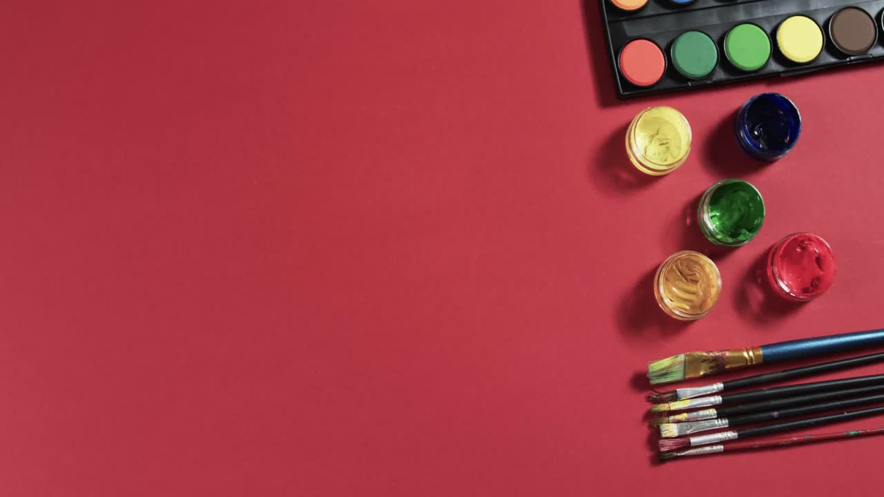 vista aérea de pinturas y pinceles dispuestos sobre un fondo rojo con espacio de copia, en cámara lenta