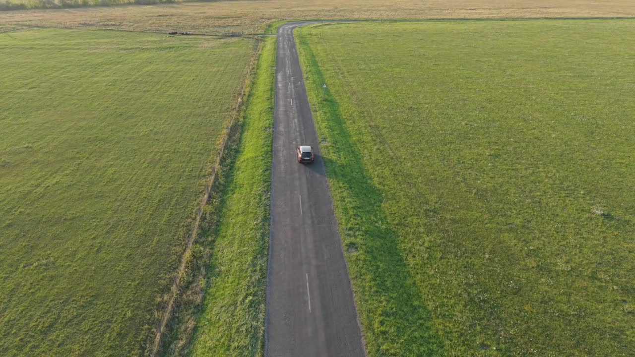 A lone car drives along an empty countryside road bordered by green fields. The peaceful rural scene captures the simplicity and tranquility of open landscapes under clear daylight