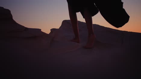 girl walking barefoot on fossil dunes wearing black abaya and ankle bracelet climbing to the top during sunset