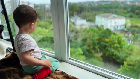 little baby boy sits on the window-sill looking outside. kid living on the upper floor looks down smiling to the camera.