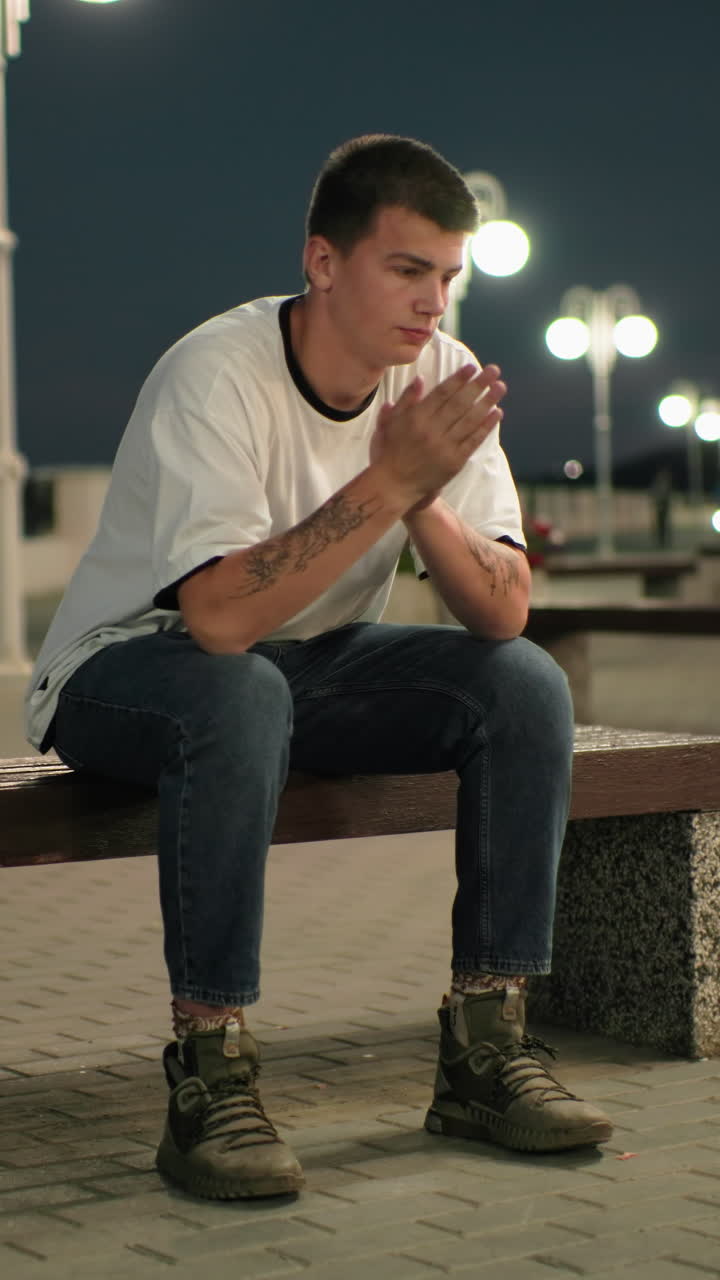 joven adulto sentado en un banco al aire libre por la noche, manos en la cara y luego quitando la mano, mirando ansioso o estresado, luces de la ciudad y peatones borrosos en el fondo
