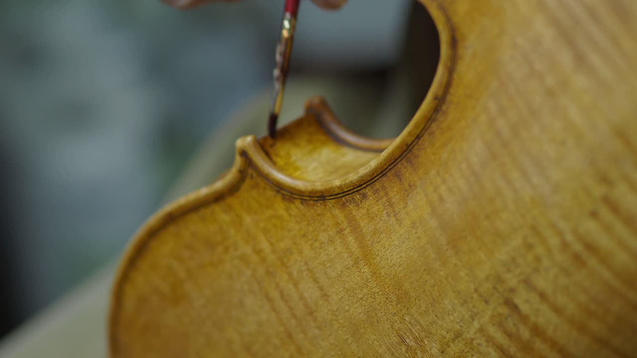 Close-up shows a luthier using a paintbrush and finger technique to apply antiquing varnish to the violin’s rib, enhancing wood texture for a classic aged appearance in a traditional workshop