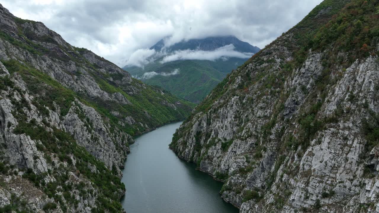 View of Lake Komani nestled between towering mountains, with a serene valley below. The dramatic cloudy mountains in the background add to the beauty and tranquility of this breathtaking landscape.