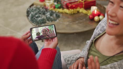 Pareja-Asiática-Sonriente-Usando-Un-Teléfono-Inteligente-Para-Una-Videollamada-Navideña-Con-Un-Hombre-Con-Sombrero-De-Papá-Noel-En-La-Pantalla