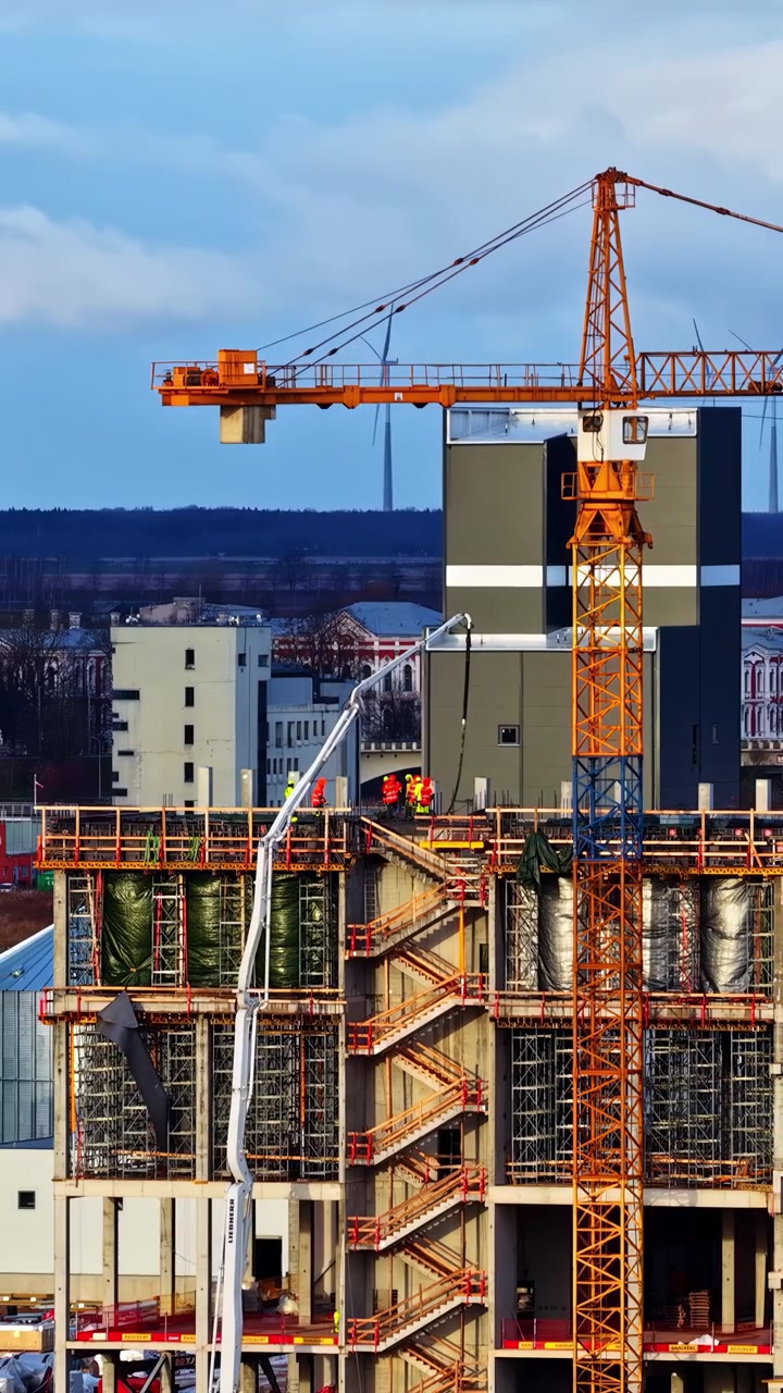 An aerial view of a busy construction site featuring a tall yellow tower crane and a white concrete pump boom working on a multistory building frame with wind turbines in the background.
