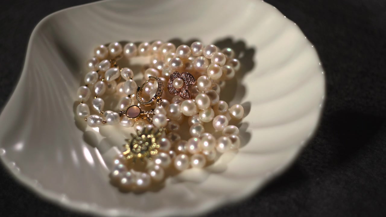 Una mano posiziona un ciondolo d'oro tra fili di perle in una ciotola di conchiglia, primo piano sull'inserimento del ciondolo, riflessi caldi