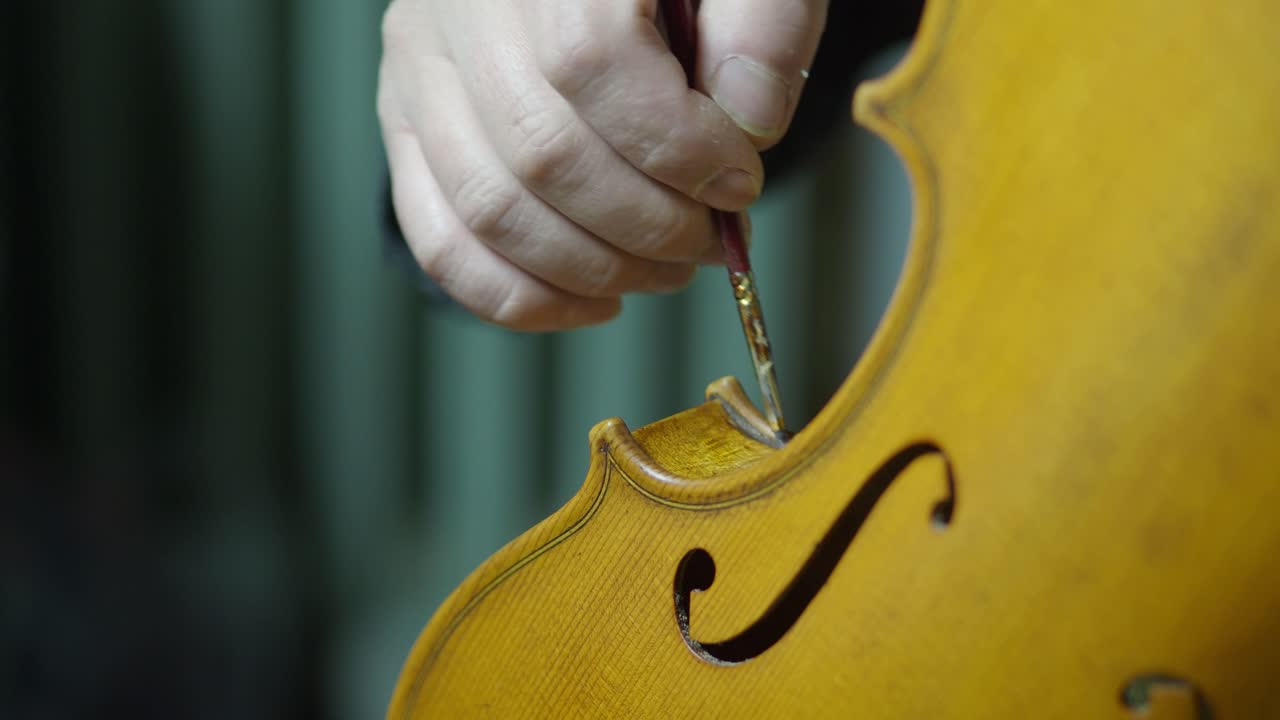Close-up of a craftsman’s hand using a fine paintbrush and finger tamponing to create antique effects on a new violin’s varnish, focusing on the center bout edges and delicate f-holes