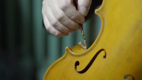 close-up of a craftsman’s hand using a fine paintbrush and finger tamponing to create antique effects on a new violin’s varnish, focusing on the center bout edges and delicate f-holes