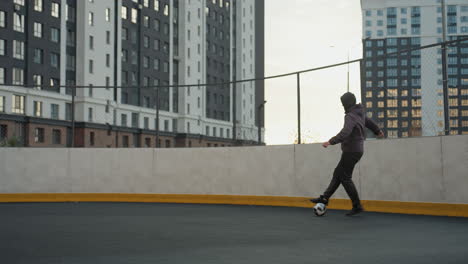 coach skillfully dribbling soccer ball on sport arena during a training session, displaying precise control and footwork, background features modern urban buildings, fencing, and soft sunlight