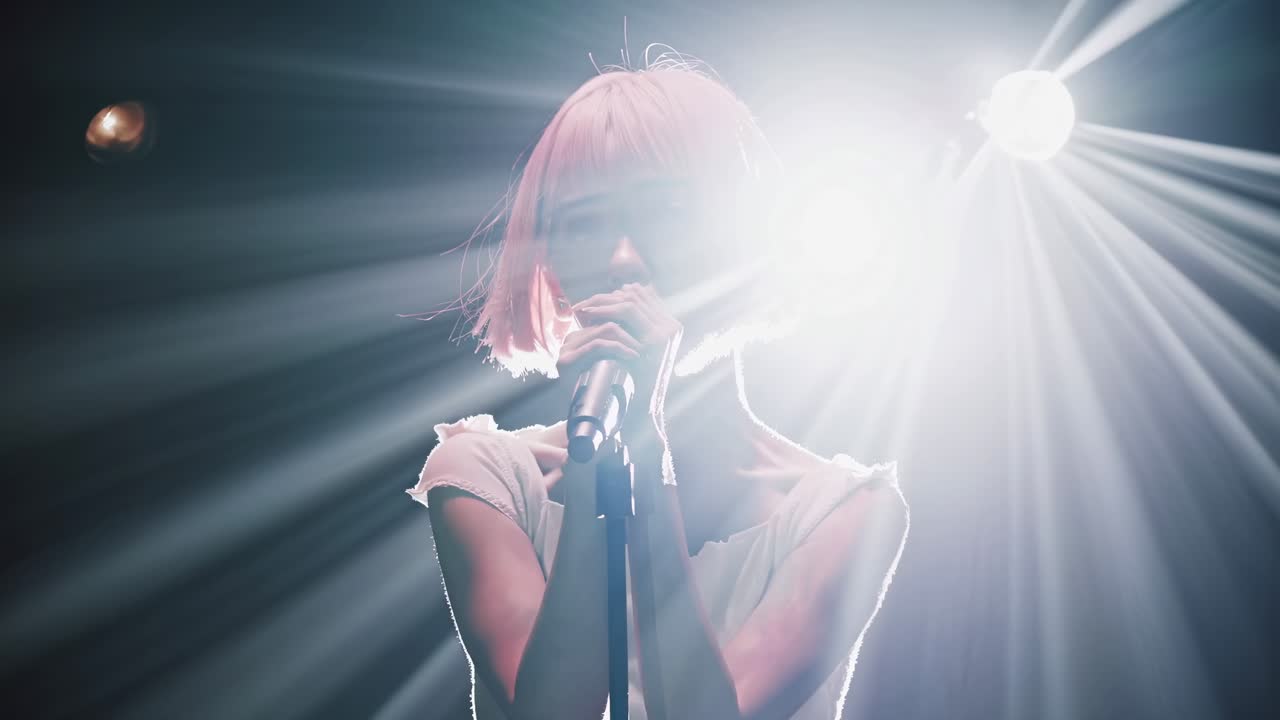 A dynamic video shot captures a singer with pink hair performing under dramatic stage lights