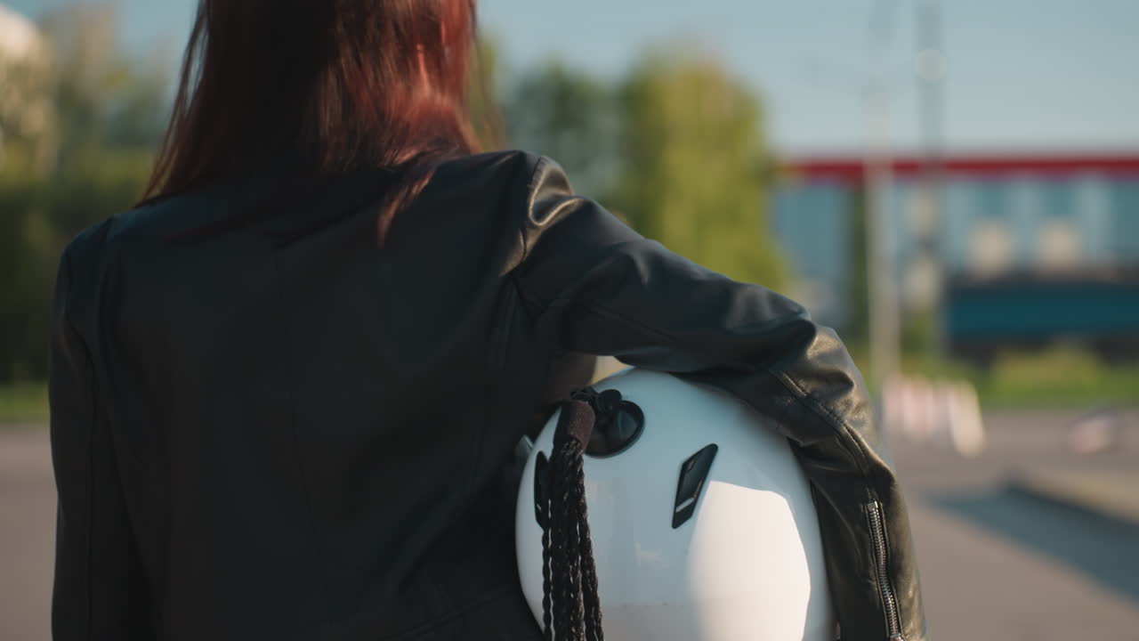 Back view of woman in black leather jacket holding white motorcycle helmet with blurred urban background, sunlight highlighting hair, creating stylish biker lifestyle