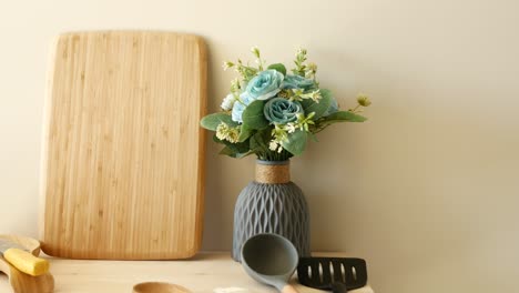 closeup of a kitchen countertop with a cutting board, wooden spoons, a spatula, a ladle, a vase with flowers and other kitchen items