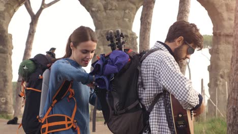 young couple backpackers tourists searching fixing open backpack near roman aqueduct arches in parco degli acquedotti park ruins in rome at sunrise with guitar and sleeping bag slow motion steadycam