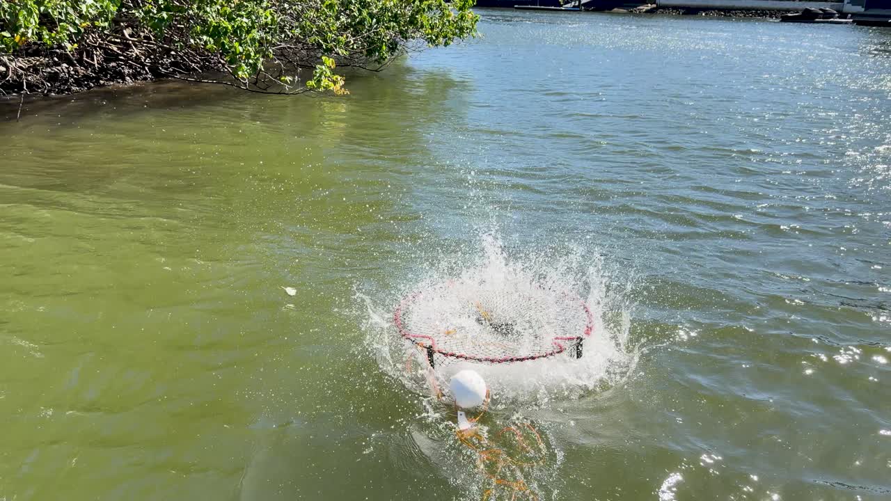 Person drops crab trap from small boat into sunlit water, slow motion, wide daylight shot