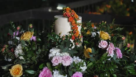 un pastel de boda blanco decorado con flores de naranja, rodeado de follaje natural y rosas blancas y rosas