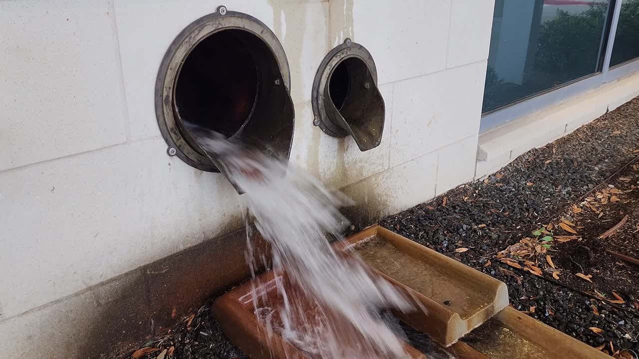 This is a short video of a large amount of water, flowing out of a drainage pipe, on the side of a building, during an afternoon thunderstorm