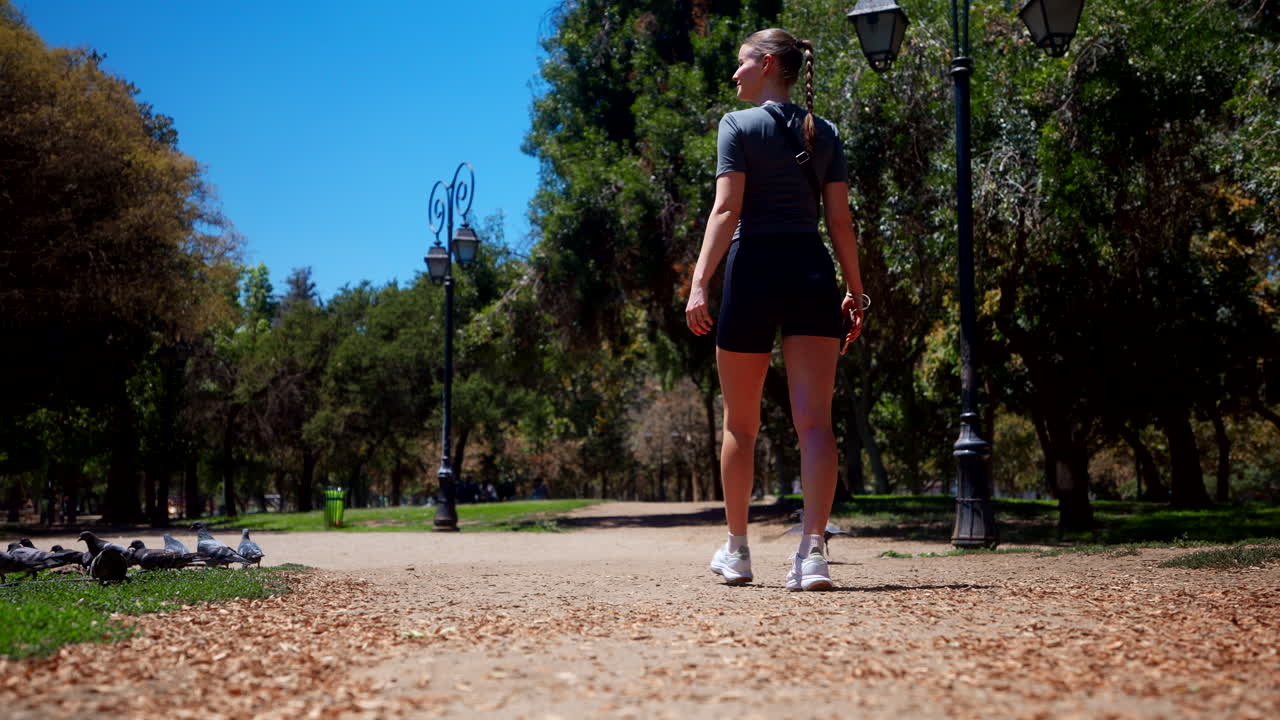 Uma jovem atraente caminha por um caminho tranquilo num parque urbano ensolarado, rodeada por árvores, pombos e postes de luz vintage num dia de verão quente e claro