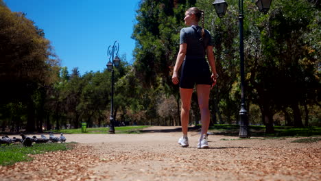 a young attractive woman walks along a peaceful path in a sunny city park, surrounded by trees, pigeons, and vintage street lamps on a warm, clear summer day
