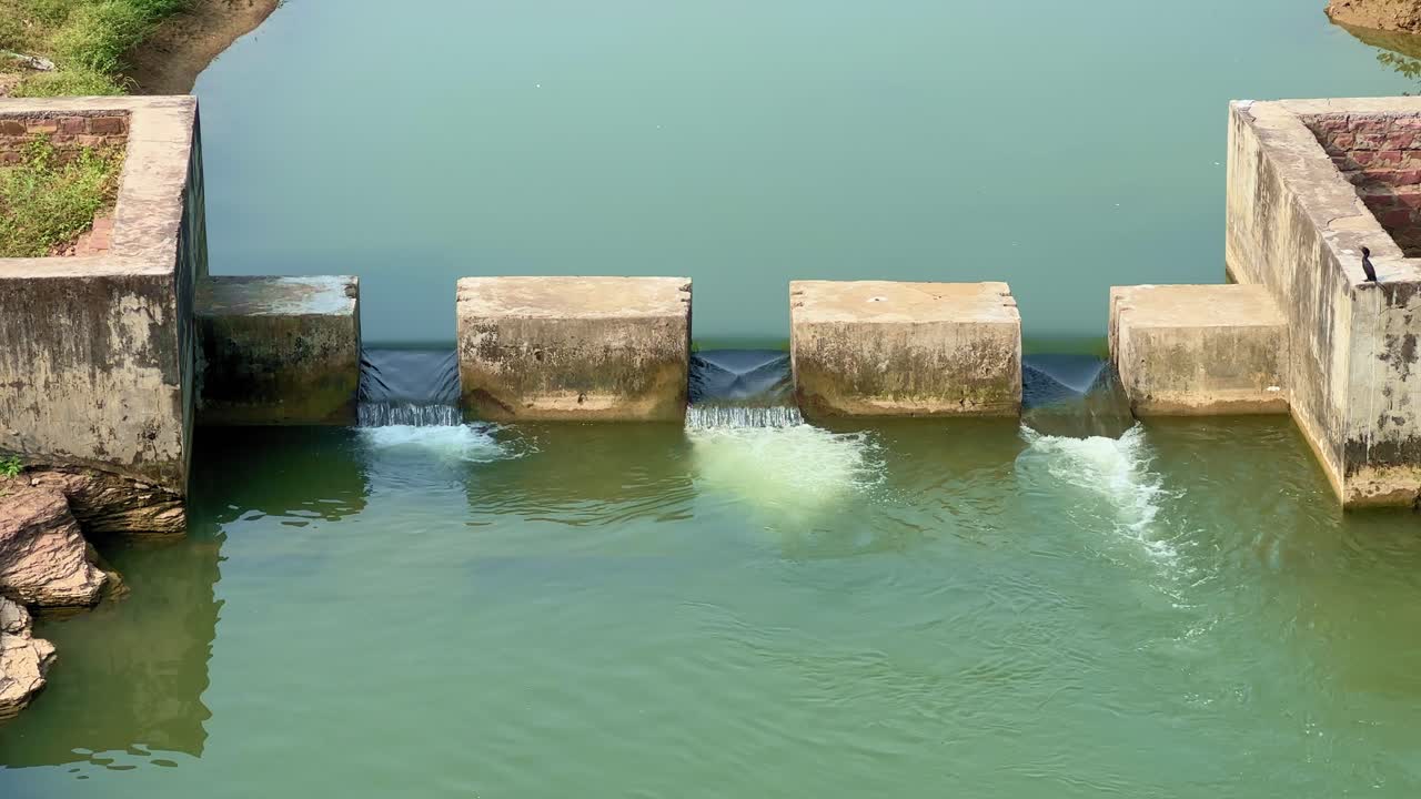 A close-up view of a small concrete check dam holding calm green water with gentle flow through narrow gates, surrounded by rocks and greenery, showcasing rural irrigation and water management
