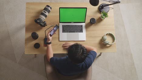 top view of a male video editor waving hand and having a video call on smartphone while using a green screen laptop next to the camera in the workspace at home