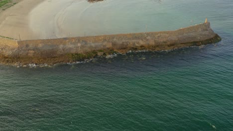 Aerial-tilt-up-from-the-vibrant-blue-waters-to-the-outer-face-of-Spiddal-Pier,-showcasing-the-pier's-structure-and-surrounding-seascape