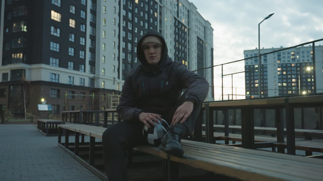 Man in hoodie sitting on bench spinning soccer ball with left hand on leg during training session, surrounded by modern residential buildings glowing with evening lights