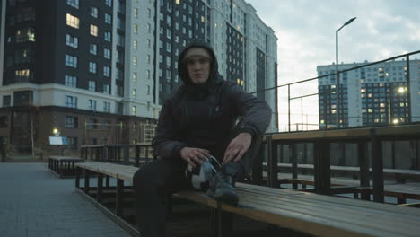 man in hoodie sitting on bench spinning soccer ball with left hand on leg during training session, surrounded by modern residential buildings glowing with evening lights