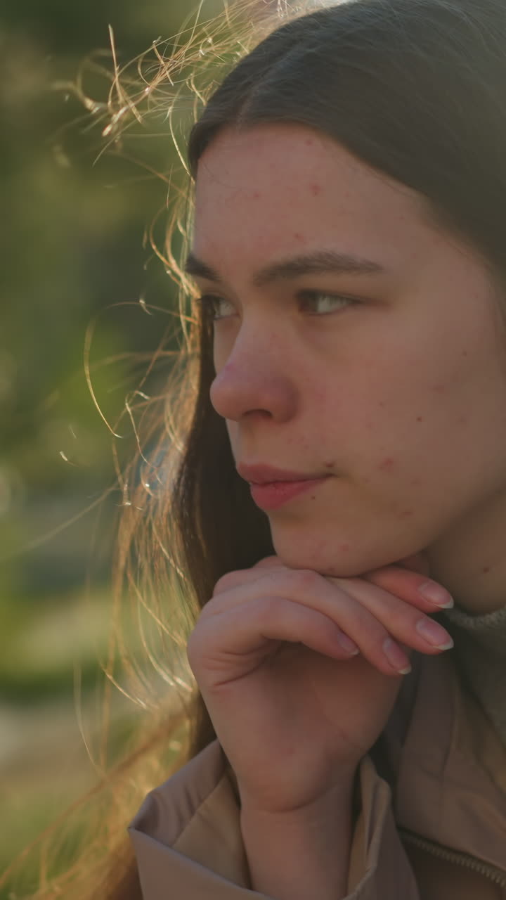 una joven con una chaqueta de melocotón está profundamente pensativa, descansando su barbilla en su mano mientras está sentada en un parque. la luz solar natural destaca su expresión pensativa contra un fondo borroso de árboles