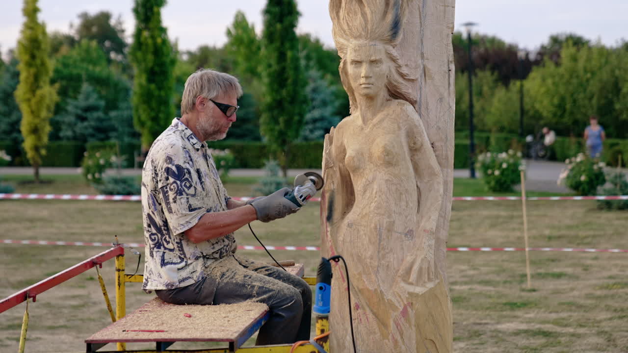 Craftsman sits outdoors working with an electric grinding instrument. Artist creating a wooden statue of a woman.