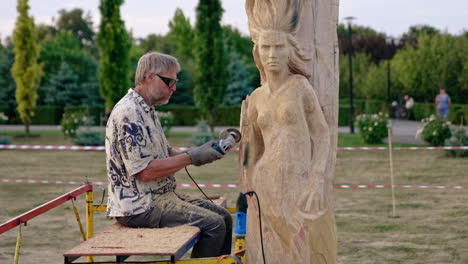craftsman sits outdoors working with an electric grinding instrument. artist creating a wooden statue of a woman.