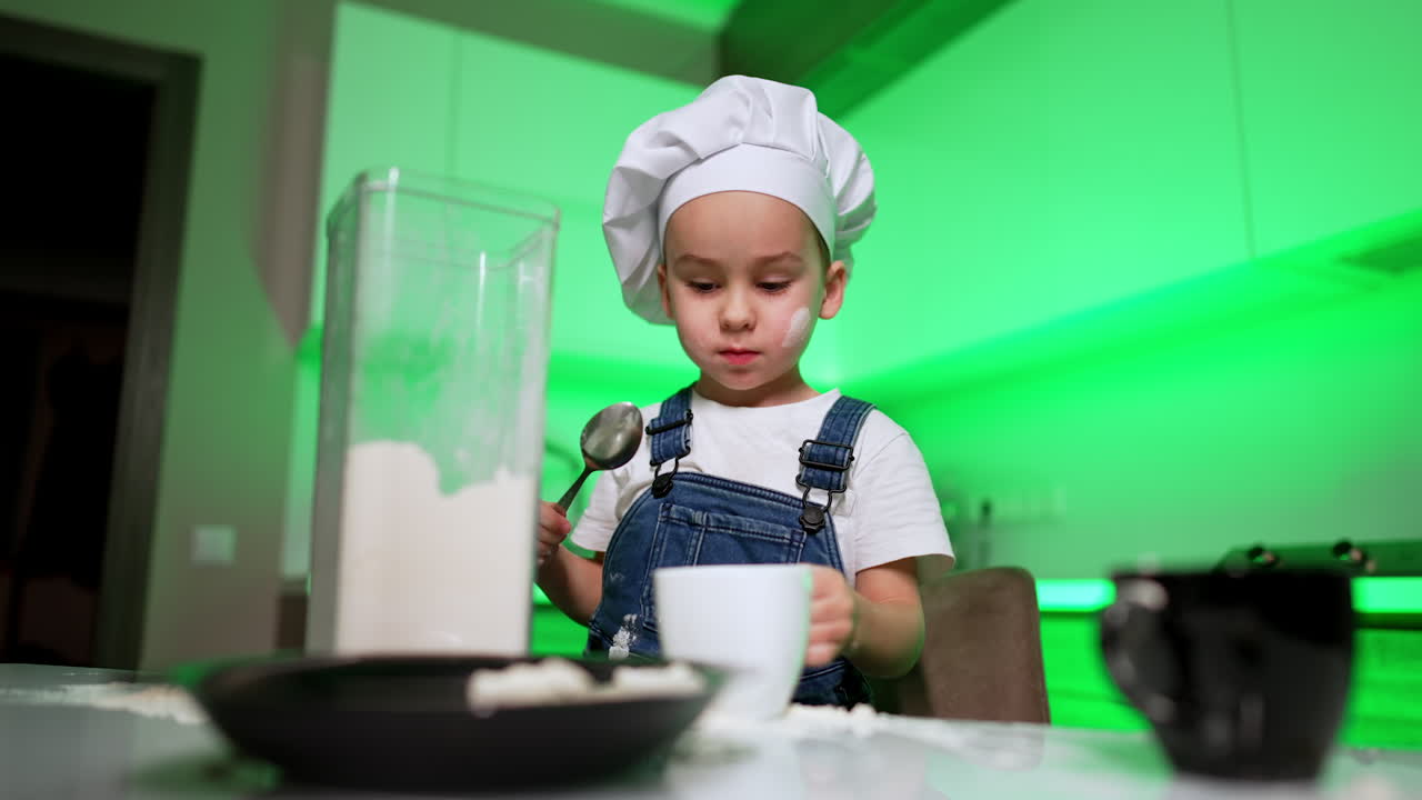 Focused Caucasian baby boy in chef's cap stands in the kitchen. Lovely toddler fills the dishes with flour. Low angle view.