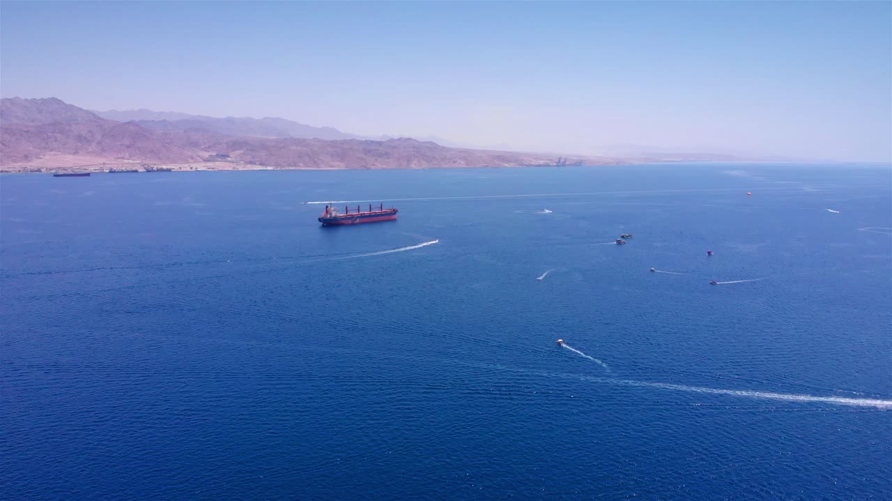 Aerial view of a cargo ship and recreational boats in a vast bay with mountains in the background