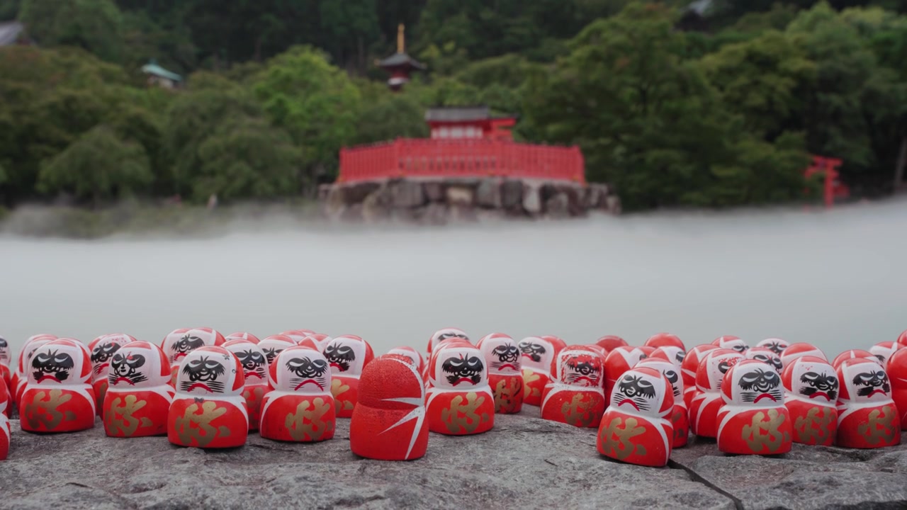 Centinaia di bambole Daruma rosse posizionate su una piattaforma di pietra al Tempio Katsuoji a Osaka, circondate da una lussureggiante vegetazione verde e una leggera nebbia, con una tradizionale struttura di tempio rosso visibile sullo sfondo