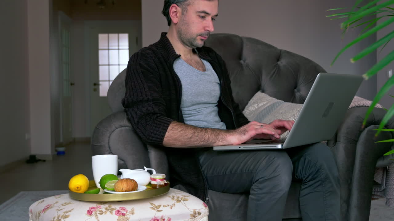 Caucasian man sitting on a chair and working on his laptop. Table with tea and fruits nearby