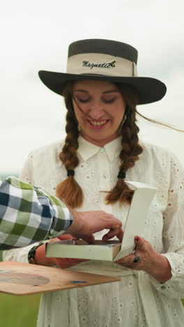 a painter wearing a black hat and plaid shirt stands in a grassy field, receiving a drawing pencil from his smiling partner. she wears a white dress and hands him the pencil with warmth and affection
