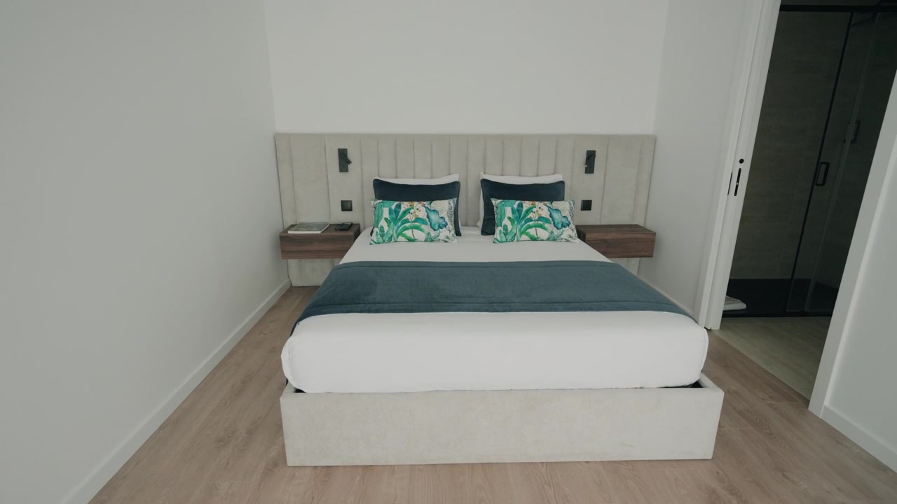 Luminosa camera da letto moderna con letto matrimoniale bianco, cuscini decorativi color verde acqua, pavimento in legno e comodini.
