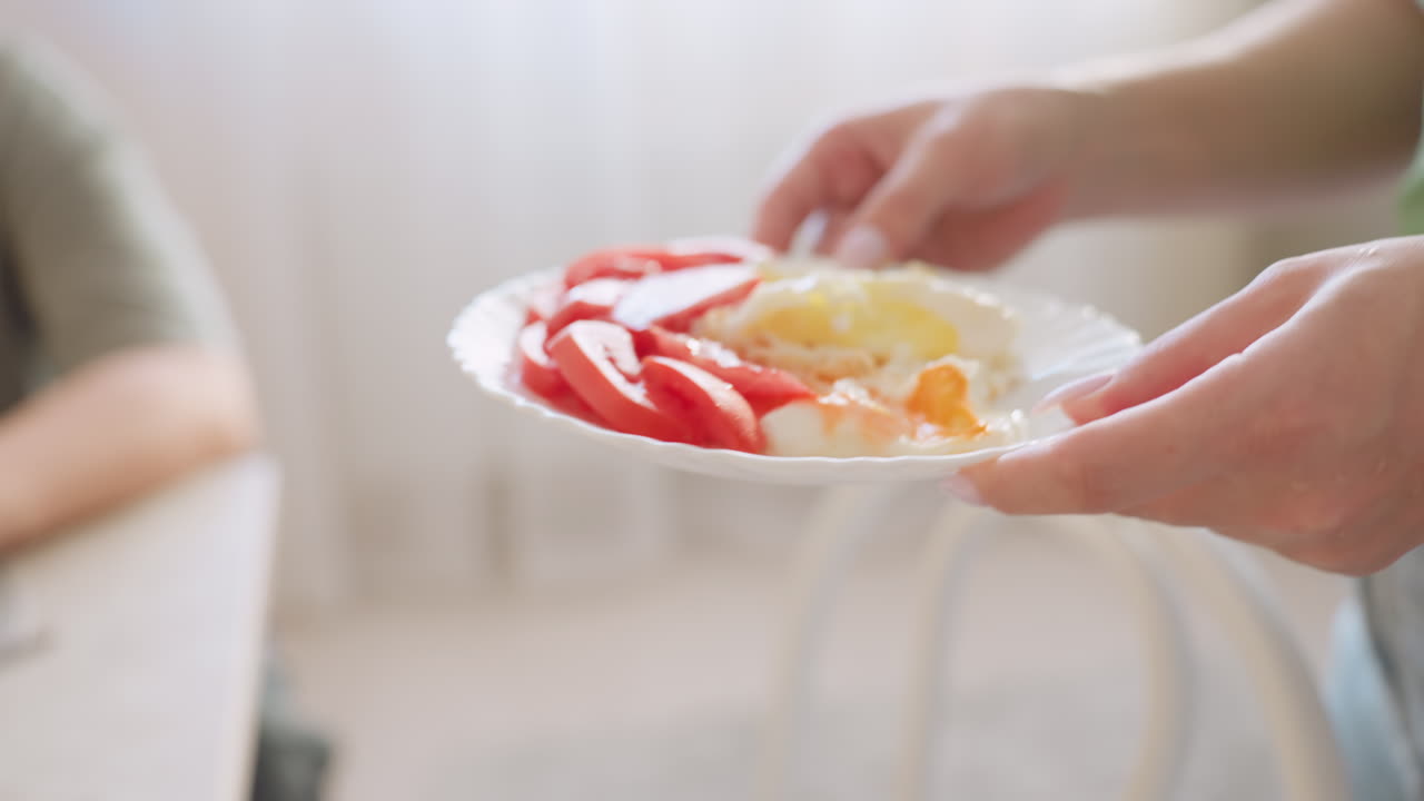 Close up of lady carrying plate with sliced tomato and fried egg to serve child seated at table, showcasing homemade breakfast preparation, healthy eating and caring household environment