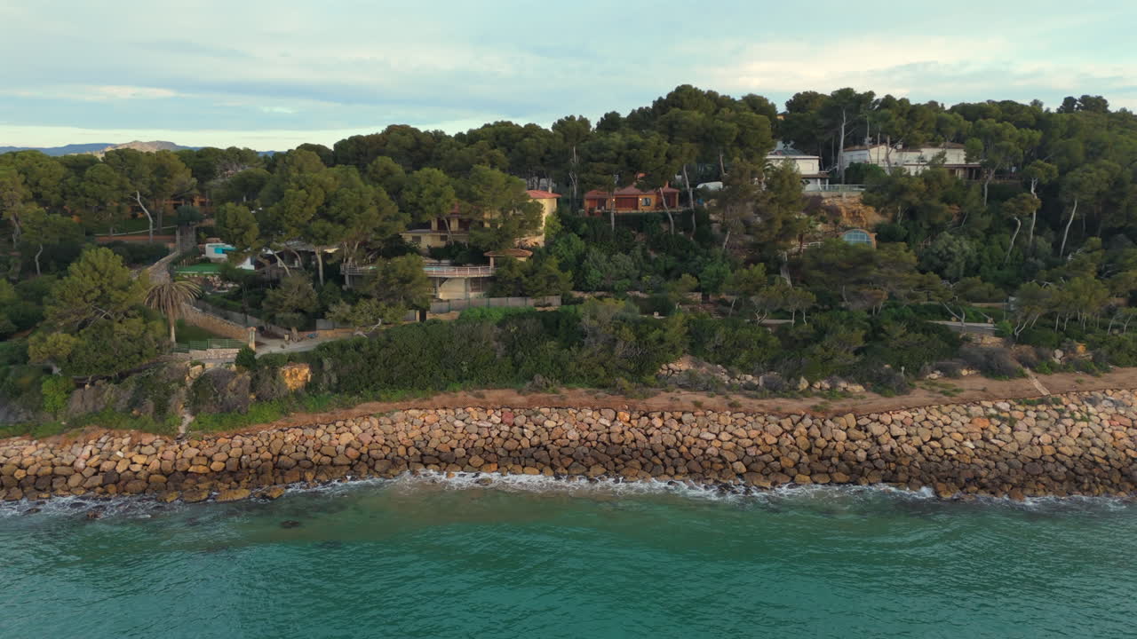 Breathtaking aerial traveling shot along a rugged Mediterranean coastline with rocky cliffs, lush greenery, and luxury villas. The turquoise sea gently crashes against the stone breakwater