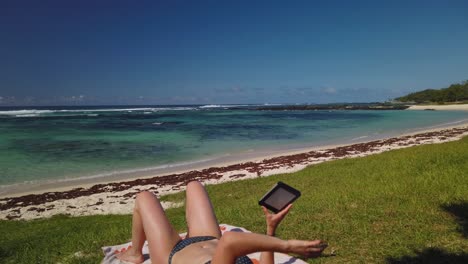 a shot of a woman reading a ebook on the grass of a tropical beach