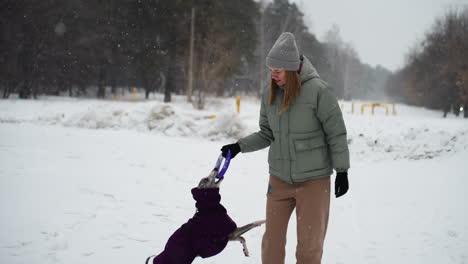 woman standing in snowy park holding toy while dog in coat eagerly looks up, anticipating playtime during cold winter day, surrounded by snow-covered landscape and bare trees in peaceful setting