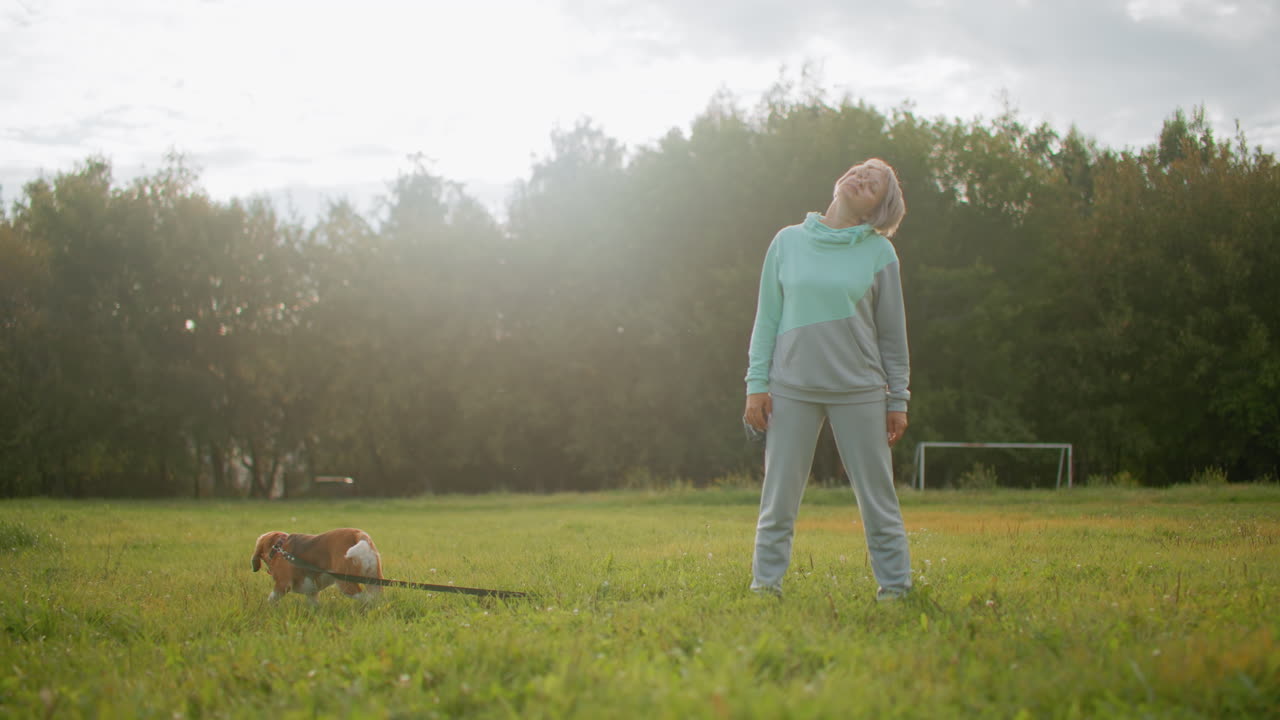 dog owner stretching neck with head tilt exercise in peaceful garden during morning light while pet dog sniffs grassy field nearby surrounded by lush green trees and calm outdoor environment