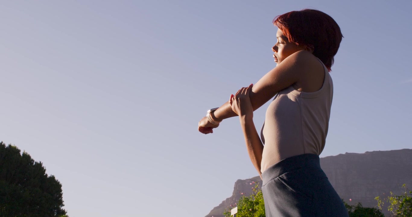 Low sun urging woman in tank-top doing arm stretches by ridge warming-up with tracker, copy space
