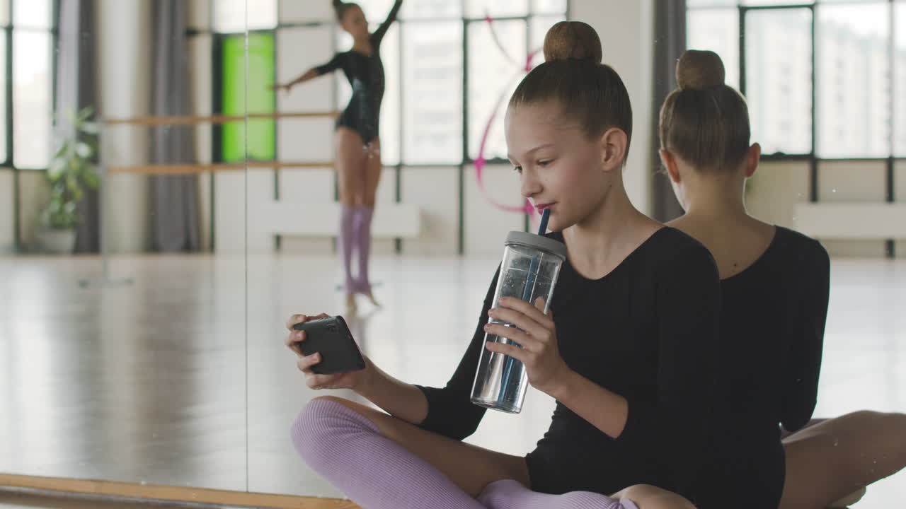 Blond gymnastikpige sidder på gulvet og tager billeder af sin veninde, der danser med et bånd